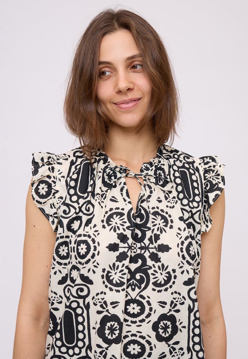Giovane donna con capelli castani di lunghezza media che indossa una blusa senza maniche a righe bianche e nere con spalline arricciate e un collo annodato.