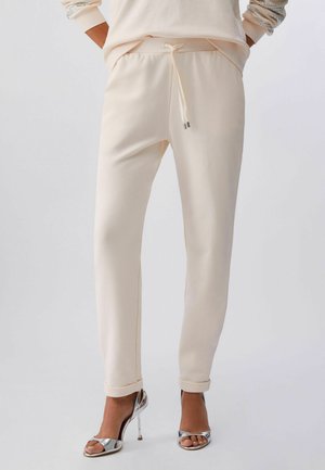 Beżowe zwężane spodnie z wiązanym w pasie sznurkiem, noszone z srebrnymi sandałami na obcasie z odkrytymi palcami na jednolitym tle.