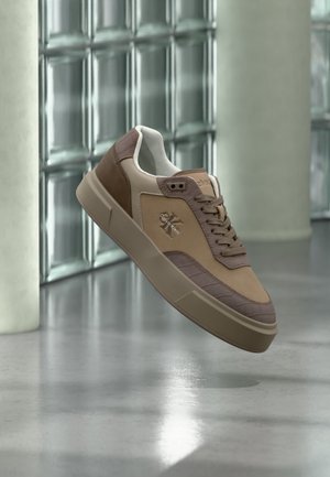 Bruine en beige Calvin Klein sneaker met veters, zwevend boven een gepolierde vloer tegen een glazen blokkenmuur.