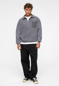 Grijze trui met een ritskraag en een opgestikte zak, gemaakt van zachte stof. Gecombineerd met zwarte, loszittende broek en zwarte sneakers.