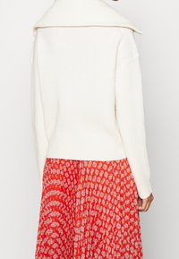 Weißer, gerippter Pullover mit weitem Kragen, kombiniert mit einem roten Rock mit wellenförmigem weißen Muster. Der Stoff wirkt weich und strukturiert.