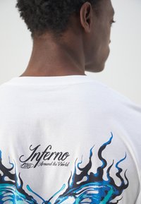 Witte katoenen t-shirt met een blauwe vlinderafbeelding en vlamontwerpen op de achterkant, met "Inferno Around the World" in zwarte tekst.