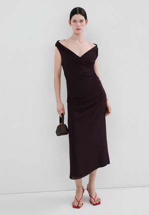 Kvinde iført en off-shoulder mørk lilla midi kjole, der holder en lille sort håndtaske og har røde sandaler med hæl og remme på.