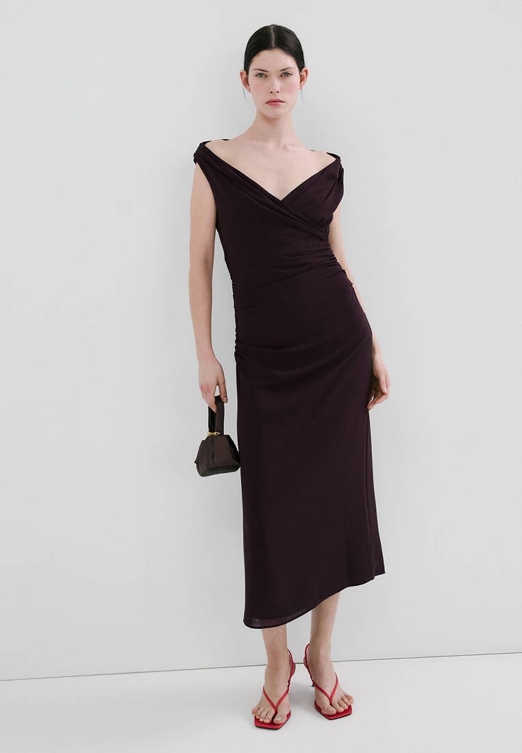 Femme portant une robe midi violette foncée décolletée aux épaules, tenant un petit sac à main noir, et portant des sandales rouges à talons avec des lanières.