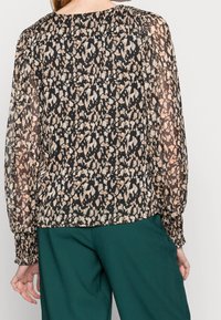 Blusa nera trasparente con una stampa astratta leopardata beige e marrone, polsini arricciati e scollo rotondo, abbinata a pantaloni verdi.