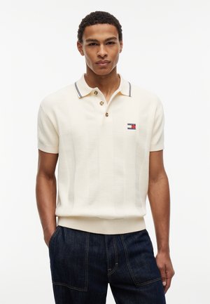 Młody mężczyzna ubrany w biały, krótki rękaw polo z guzikami i małym prostokątnym logo, w połączeniu z ciemnoniebieskimi dżinsami.