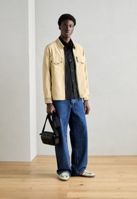Beige jas met twee voorzakken, zwart overhemd met knopen, blauwe wijde jeans en groen-witte sneakers, houdt een zwarte tas vast.