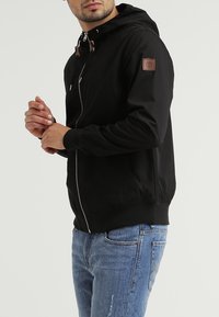 Czarna rozpinana na zamek kurtka z kapturem wykonana z gładkiego materiału. Posiada brązową naszywkę z logo oraz elastyczne mankiety, zestawiona z niebieskimi, postarzanymi dżinsami.