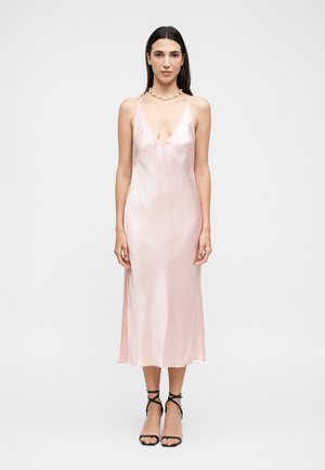 Kvinde iført en sart lyserød satin natkjole med tynde stropper, V-udskæring, sorte remsko med hæle og en perlebesat halskæde, stående mod en ensfarvet baggrund.