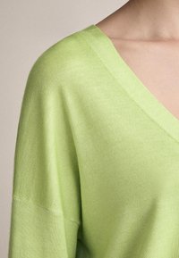 Maglione verde chiaro con scollo a V e maniche corte. Il tessuto ha una texture liscia, mostrando un design semplice e moderno.