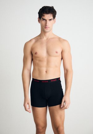 Schwarze Boxershorts mit einem roten "HUGO"-Bund, enganliegendem Design, weicher Textur und mittellanger Beinlänge, getragen von einem oberkörperfreien männlichen Modell.