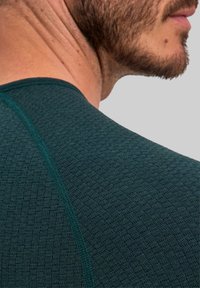 Ciemnozielona sportowa koszulka z teksturowaną tkaniną o subtelnym wzorze, zaokrąglonym dekoltem i kontrastowymi przeszyciami.