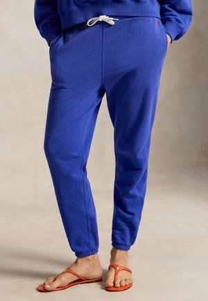 Személy kék futónadrágot visel fehér zsinórral és narancssárga papucsban, kezét a zsebébe dugva, egyszínű háttér előtt áll.