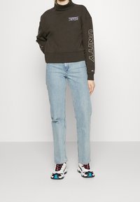Persona che indossa una felpa nera Tommy Jeans con "UNITY" sulla manica, jeans azzurri a gamba dritta e scarpe da ginnastica bianche con accenti blu.