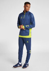 Marinträningsoverall med limegröna detaljer, tillverkad av slätt, stretchigt material; har en halvmatchande zip-tröja med logotyp och chevrondetaljer på byxorna.