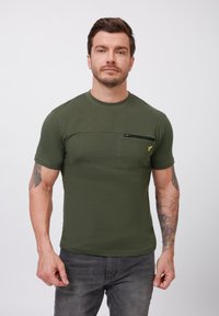 Olivengrøn kortærmet t-shirt med rund halsudskæring, prydet med en påsyet lomme og en kontrasterende lynlåsdetalje; stoffet virker glat og let.