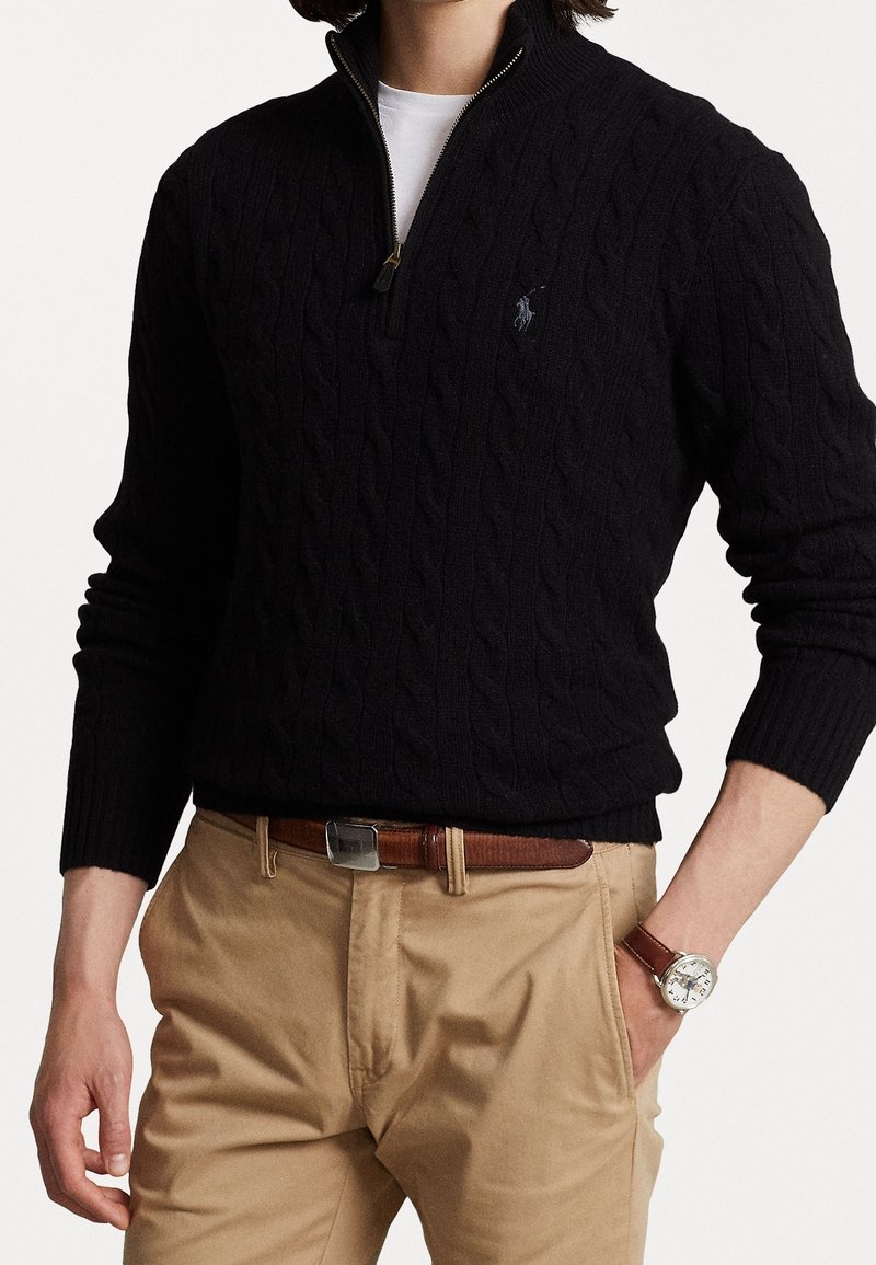 Personne portant un pull noir torsadé à col zippé sur une chemise blanche, un pantalon beige avec une ceinture marron et une montre-bracelet argentée.