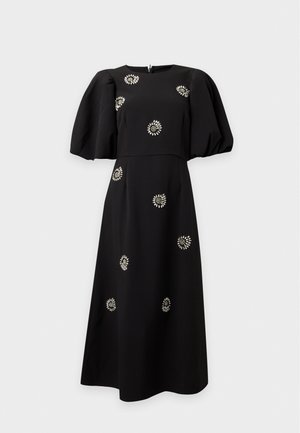 Femme aux longs cheveux raides portant une robe midi noire avec des manches bouffantes et des ornements floraux en perles, debout avec une main dans la poche.