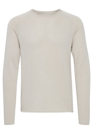 Man wearing cream long-sleeve sweater, olive green jogger pants, and white sneakers, standing against plain light background.