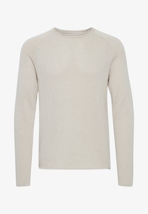 Homme portant un pull crème à manches longues, un pantalon de jogging vert olive et des baskets blanches, debout devant un fond clair uni.