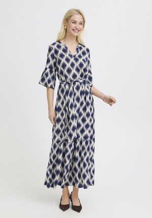 Blonde vrouw die een midi-jurk met ceintuur draagt, blauw-wit patroon, met halve mouwen en zwarte puntige hakken, glimlachend tegen een effen achtergrond.