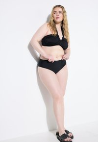 Černý bikini s topem ve stylu bandeau s twistem a vysokými kalhotkami. Hladký materiál, minimalistický design a nošeno s černými skládacími sandály.