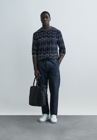 Tamsiai mėlynas megztinis su raštu, laisvo silueto, derinamas su tamsiomis siaurėjančiomis kelnėmis ir baltais sportbačiais. Nešiojama juoda siūta rankinė.