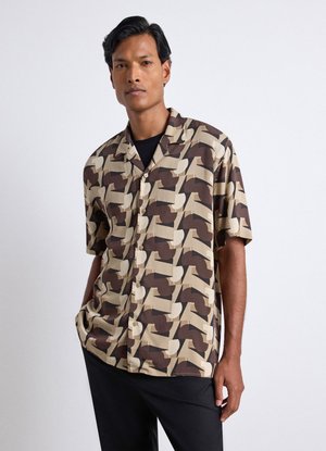 Uomo che indossa una camicia a maniche corte con motivo geometrico astratto beige e marrone scuro, in piedi con una mano in tasca.