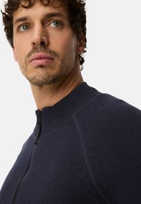 Navy zip-up sweater with a high ribbed collar, textured knit pattern on shoulders, and sleek black zipper detail.