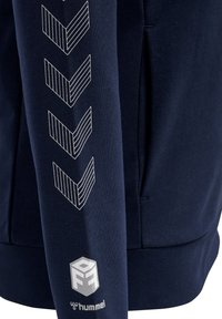 Marinblå hoodie med vit chevron grafik på ärmen och logotyp vid fållen. Tillverkad av mjukt material med en slät textur.