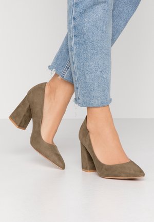 Fødder iført lyseblå jeans med frynset kant og taupe ruskindspumps med blokhæl på en hvid gulv mod en ensfarvet baggrund.