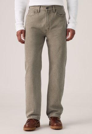 Mann trägt hellbraune Jeans mit geradem Bein, ein weißes, geripptes Langarmshirt und braune Lederschuhe mit robuster Sohle.