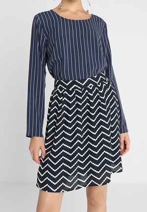 Frau trägt ein marineblaues Top mit weißen vertikalen Streifen und einen marineblauen Rock mit weißen Zickzackmustern, stehend vor einem einfarbigen Hintergrund.