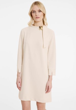 Cremfarbiges Kleid mit hohem Kragen, langen Ärmeln und einer Schleifendetail am Kragen. Akzentuiert durch ein Hardware-Teil in Goldfarbe. Glattes Material.