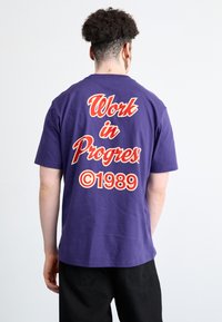 Purple cotton t-shirt with "Work in Progress" printed in red and white on the back, featuring ©1989 in a circular design below the text.