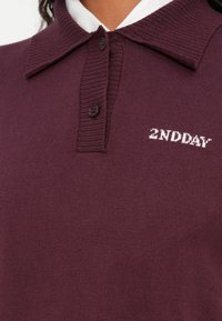Bordo pulover s rebrastim ovratnikom, dvostrukim gumbom i izvezenim logotipom "2NDDAY" u bijeloj boji na prsima. Mekana pletena tekstura, ležeran dizajn.