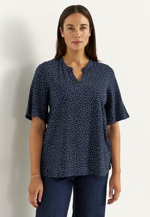 Vrouw met lang donker haar die een marineblauw blouse met korte mouwen en witte polkadots draagt, en een donkere blauwe spijkerbroek, staand tegen een effen achtergrond.