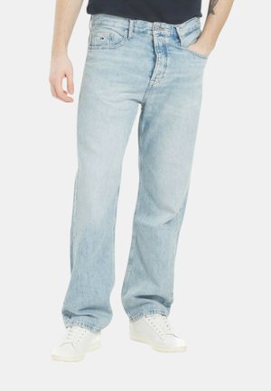 Uomo che indossa jeans azzurri a gamba dritta e sneakers bianche, con una mano in tasca su sfondo uniforme.