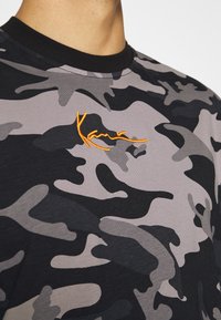 Černé a šedé maskáčové tričko s kulatým výstřihem, zdobené oranžovým vyšitým logem. Měkká, texturovaná látka s moderním designem.
