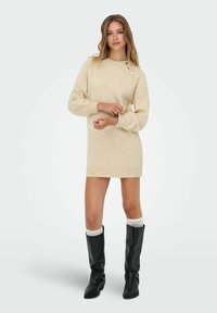 Beige trui jurk in een zachte textuur met een ronde halslijn, voorzien van vier decoratieve knopen op één schouder en geribbelde zoomranden.