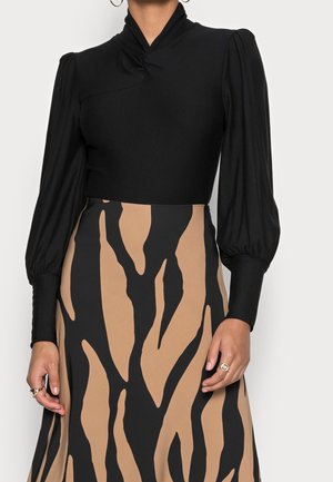 Frau trägt ein schwarzes Langarmoberteil mit Puffärmeln und hohem Halsausschnitt, kombiniert mit einem beigen und schwarzen Rock mit abstraktem Muster.