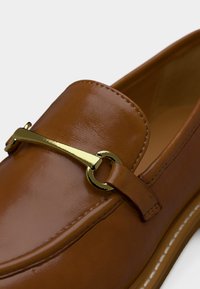 Καφέ δερμάτινο μοκασίνι με χρυσή μεταλλική λεπτομέρεια σε σχήμα άλογου και εμφανή ραφές κατά μήκος της άκρης της λαστιχένιας σόλας σε λευκό φόντο.