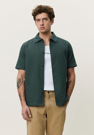 Junger Mann mit lockigem Haar, der ein strukturiertes dunkelgrünes Kurzarmhemd über einem weißen Grafik-T-Shirt und beigen Hosen trägt, steht vor einem schlichten Hintergrund.