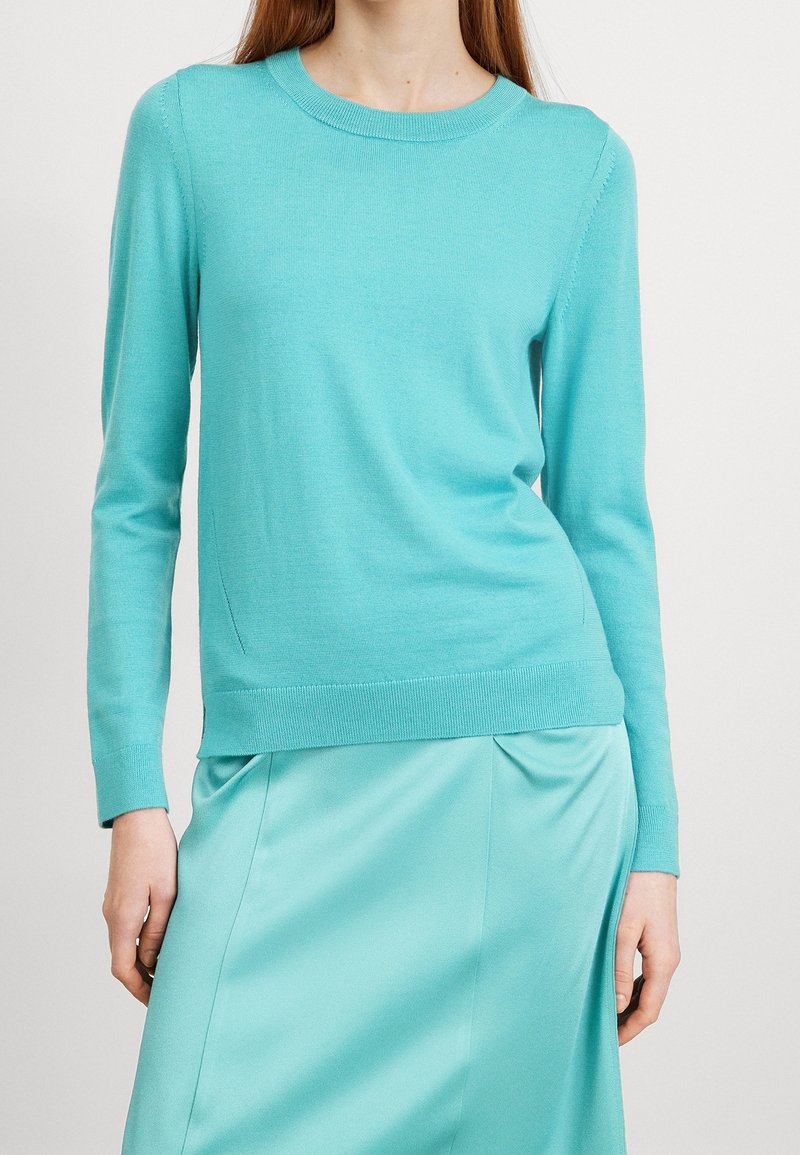 Pull ajusté à manches longues turquoise assorti à une jupe en satin turquoise clair portée par une personne sur un fond uni.