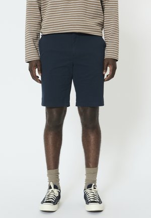 Laivastonsiniset shortsit suoralla leikkauksella, yhdistetty raidalliseen pitkähihainen paitaan. Mustat tennarit valkoisilla yksityiskohdilla ja vaaleanruskeat sukat.