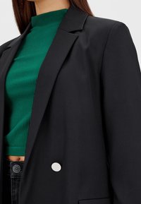 Blazer nero con rever a punta, dettaglio a singolo bottone argento, indossato sopra un dolcevita verde a coste. Contrasto di texture nei materiali.