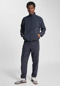 Marinblå sportsoverall med zip-jacka och byxor med dragsnöre. Utrustad med vit kantning, elastiska muddar och sidofickor. Matchas med grå sneakers.