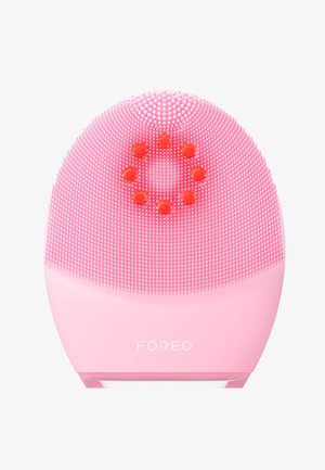 Pink facial cleansing device with textured silicone bristles, circular accent feature, and a rounded shape. Logo displayed on the base.