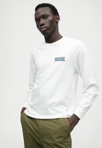 Langärmliges weißes T-Shirt aus Baumwolle, mit einem bunten "RIPCURL"-Logo auf der Brust. Glatte Textur, entspannter Schnitt.