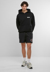 Hanorac negru cu buzunar frontal și logo alb, asortat cu pantaloni scurți negri cu un grafic circular roșu și alb. Adidași gri și șosete albe.